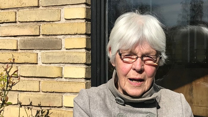 En kvinde med hvid hår og briller står foran en mur af gule teglsten.