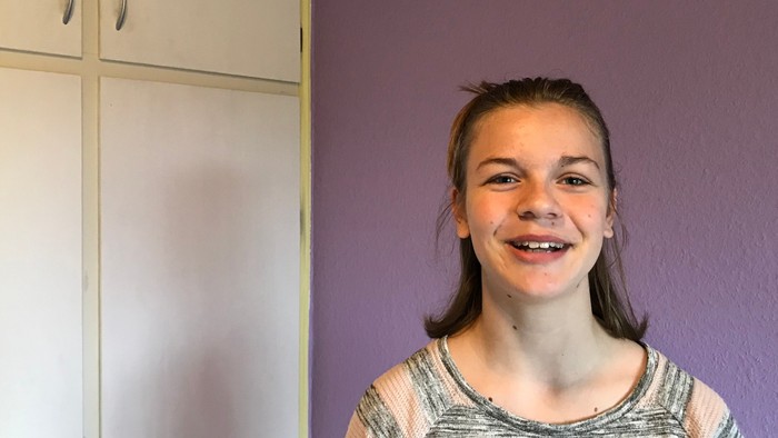 En pige med langt brunt hår står foran en lilla væg og en hvid køleskab.