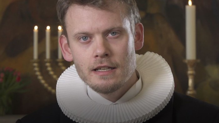 En mand med hvid krage og sort klæde står foran en lysestake.