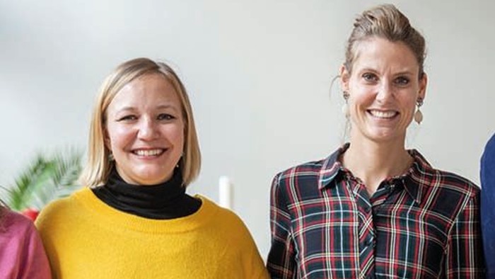 Zwei Frauen lächeln in die Kamera, eine trägt ein gelbes Pullover, die andere ein kariertes Kleid