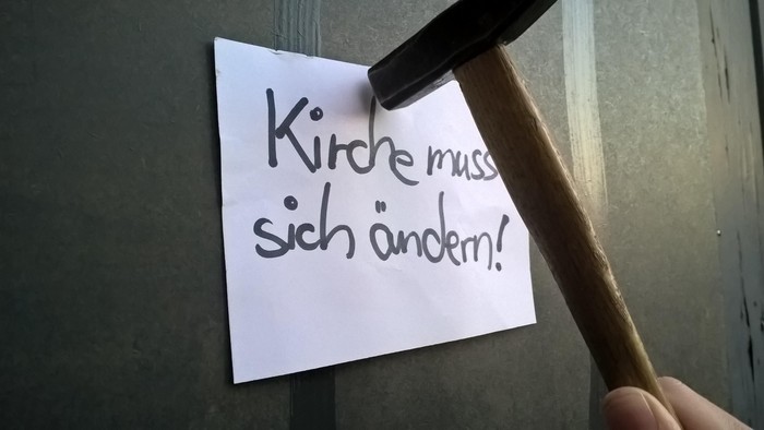 Eine Hand hält einen Hammer über einen Zettel mit der Aufschrift *„Kirche muss sich ändern!“*.