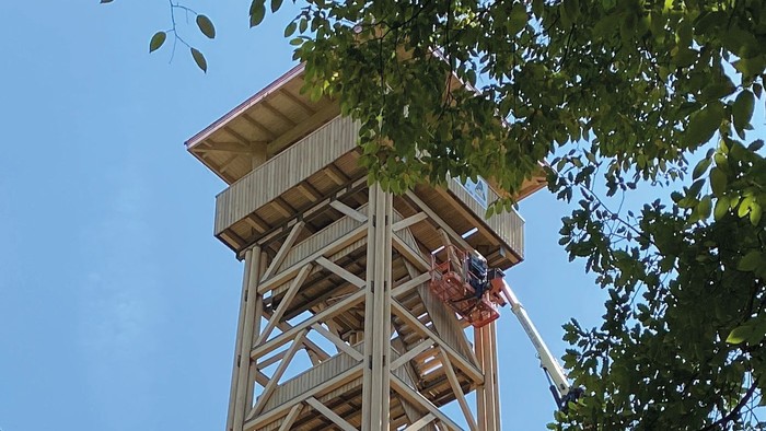 Holzturm mit Leiter und Menschen, umgeben von Bäumen und blauen Himmel