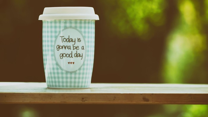 Eine grüne Kaffeetasse mit einem positiven Spruch steht auf einem Holzbalkon.