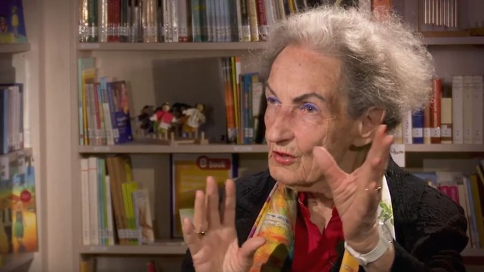 Eine ältere Frau gestikuliert lebhaft vor Bücherregalen.