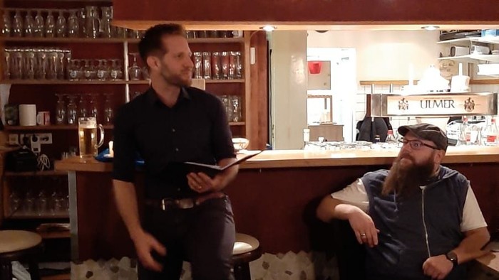 Zwei Männer sitzen in einem Café, einer steht und liest ein Buch.