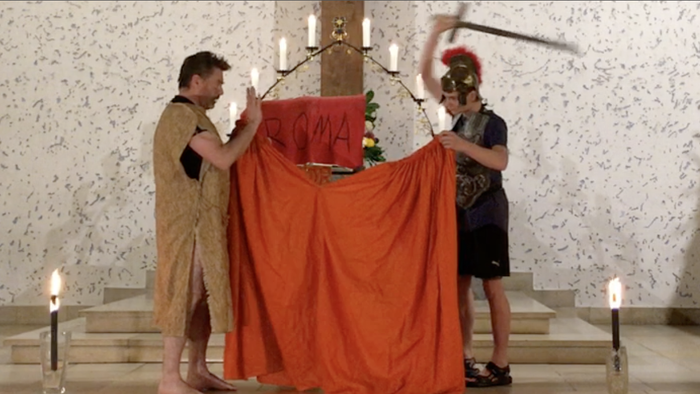 Zwei Personen heben ein großes orangefarbenes Tuch auf einer Bühne.