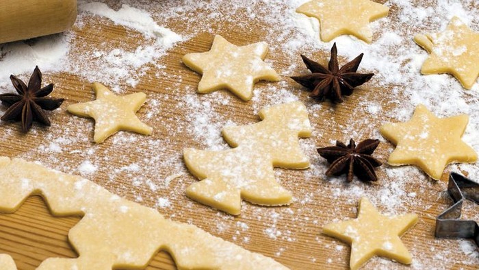 Selbstgemachte sternförmige Zuckerplätzchen, mit Mehl bestäubt, auf einer Holzfläche mit Backutensilien.