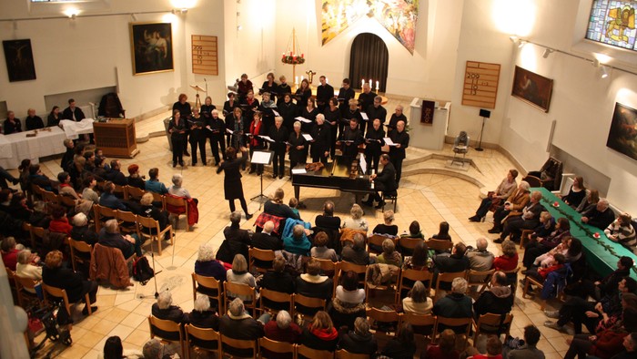 Eine Gruppe von Menschen singt in einem großen Raum mit hohen Decken und Buntglasfenstern, während eine Menge auf Stühlen sitzt und zusieht.