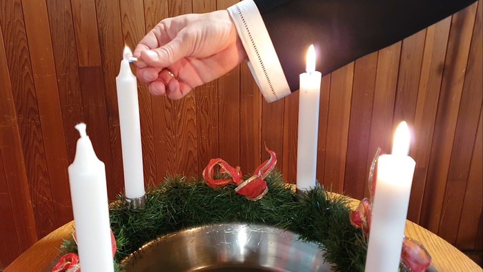 Hand lighting white candle on wreath with red ribbon and other lit candles.