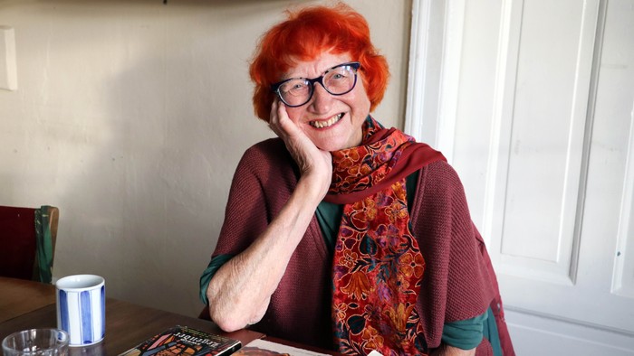 En kvinde med rødt hår og briller sitter ved et bord med bøger og en tærte.