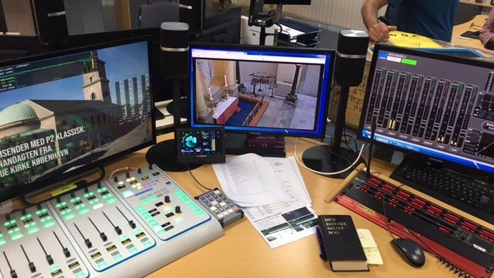 Bureau med tre skærme, tastatur, mus og lydudstyr. En person sitter til højre.