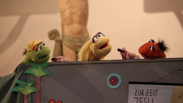 Drei Puppen sitzen auf einer Bühne, eine hält ein Mikrofon, eine andere ein Schild mit 
