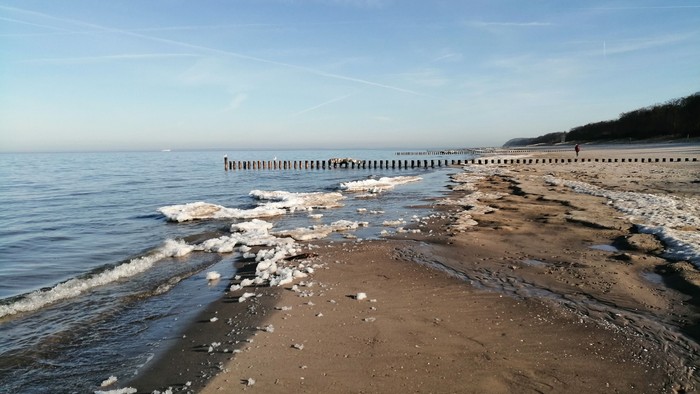 Ruhige Strandküste mit sanften Wellen, die auf eisige Stellen im Sand treffen, unter klarem Himmel.