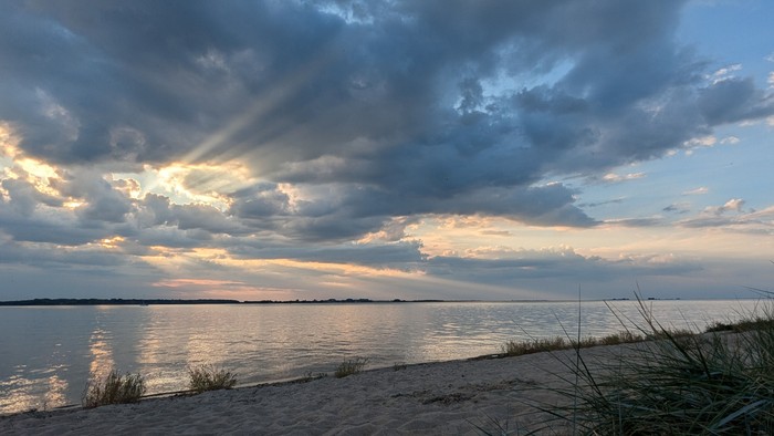 Ruhige Uferlinie bei Dämmerung mit dramatischen Wolken, die sich im stillen, schimmernden Wasser spiegeln.
