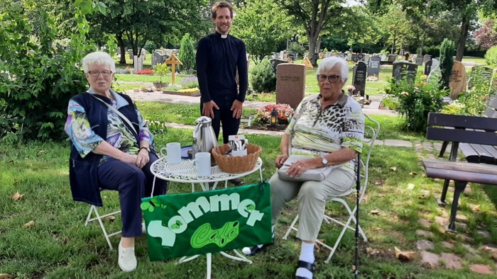 Drei Personen sitzen im Park bei einem Picknick, eine grüne Tischdecke mit 