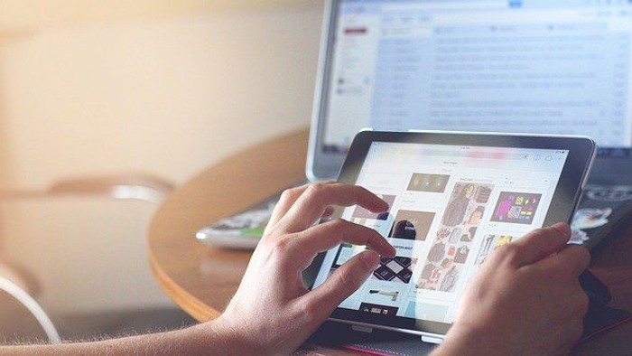 Person, die auf einem Tablet Kleidungsoptionen durchstöbert, während im Hintergrund ein Laptop läuft.