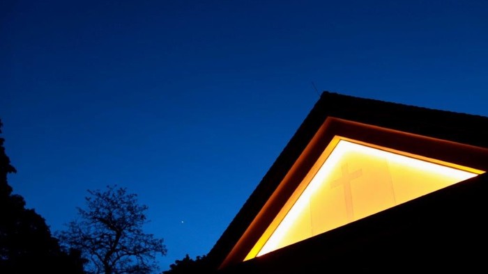 Ein Haus mit beleuchtetem Dachfenster vor dunklem Himmel und Bäumen.