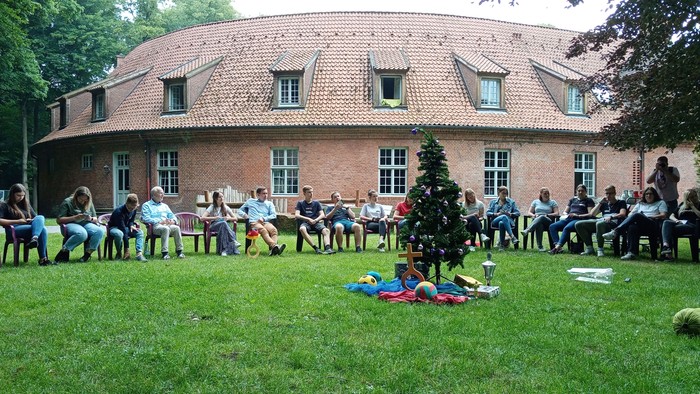 Gruppe von Menschen sitzt vor einem Haus auf Stühlen und hört einem Redner zu
