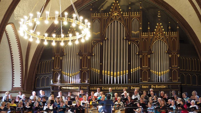 Ein Chor singt in einer großen, prächtigen Kirche mit einem großen Orgelpfeifenwerk und einem großen Kronleuchter.