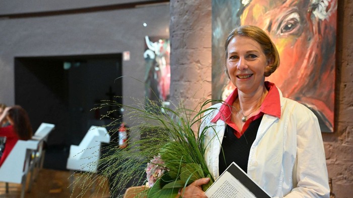 Eine Frau im weißen Kittel hält Blumen und ein Buch, lächelt in einer Galerie mit Kunstwerken im Hintergrund.