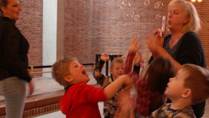 Børn danser og spiller i et rum med mur af ruder og hængelys.