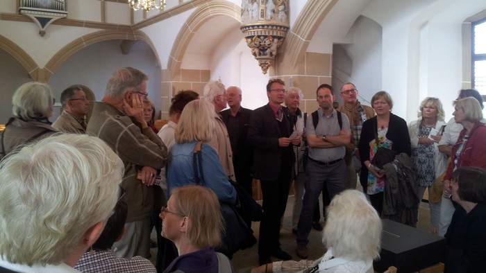 Eine Gruppe von Menschen versammelt sich in einem großen, hellen Raum mit hohen Decken und einem Kronleuchter.