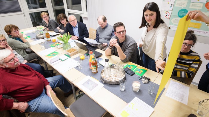 Eine Gruppe von Menschen sitzt an einem Tisch und arbeitet an einem Projekt, es gibt Papiere, Stifte und ein gelbes Schild.