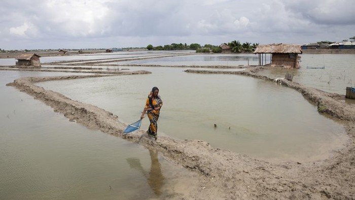 Eine Person trägt einen Eimer durch ein flaches Wassergebiet mit kleinen Hütten im Hintergrund