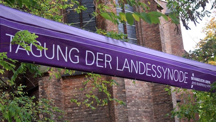 Purpurfarbiges Schild mit deutscher Aufschrift vor Ziegelmauer und Grünpflanzen