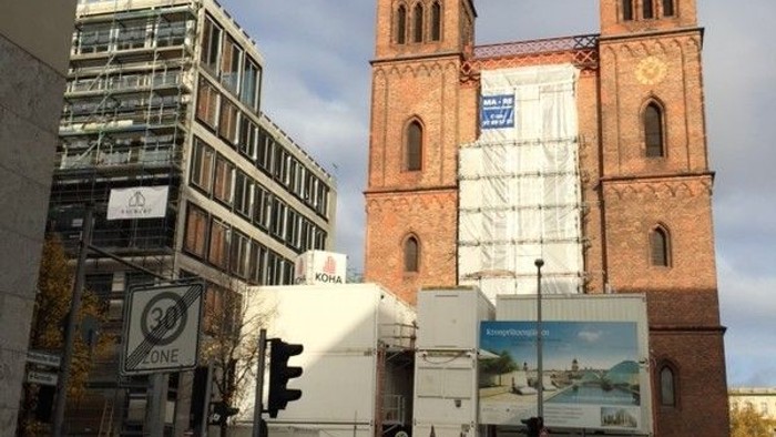 Altes Backsteingebäude mit zwei Türmen wird saniert, umgeben von modernen Bürogebäuden und Baustellen.