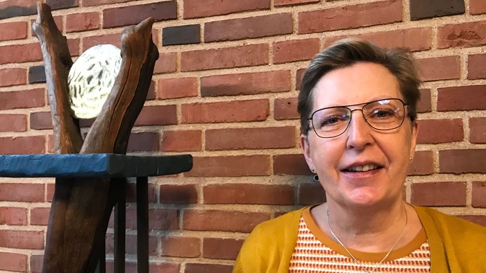 Kvinde med briller og gult skærmtøj står for en mur med urolige klokker.