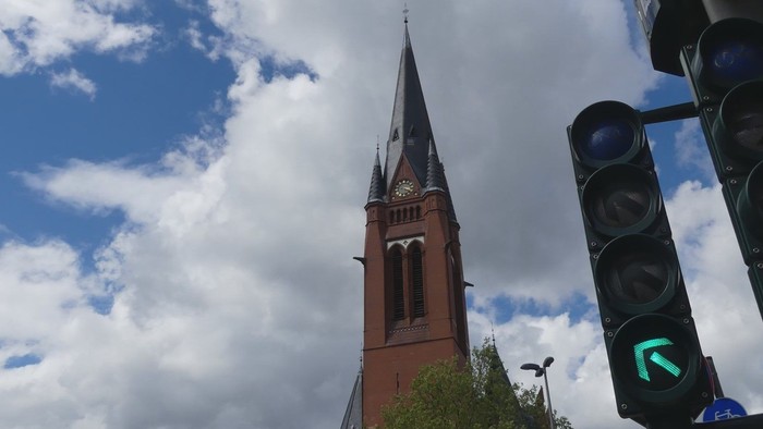 Ein roter Kirchturm mit spitzer Spitze vor blauem Himmel und grünen Ampeln.