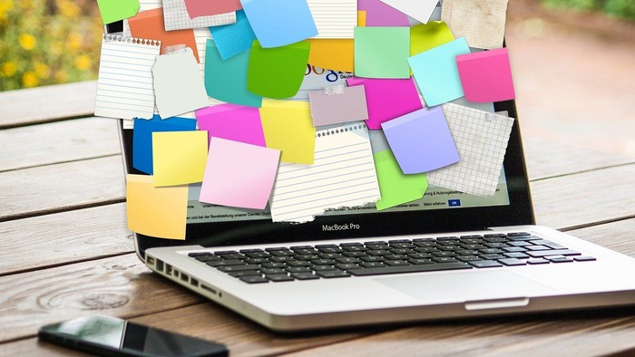 En MacBook Pro dekket af mange farvede post-it noter, placeret på en træbæn ved siden af et mobiltelefon.