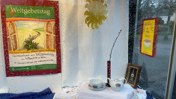 Ein Fenster mit Tee-Set, Obst und Dekorationen, daneben ein Poster mit "Weltgebetstag"