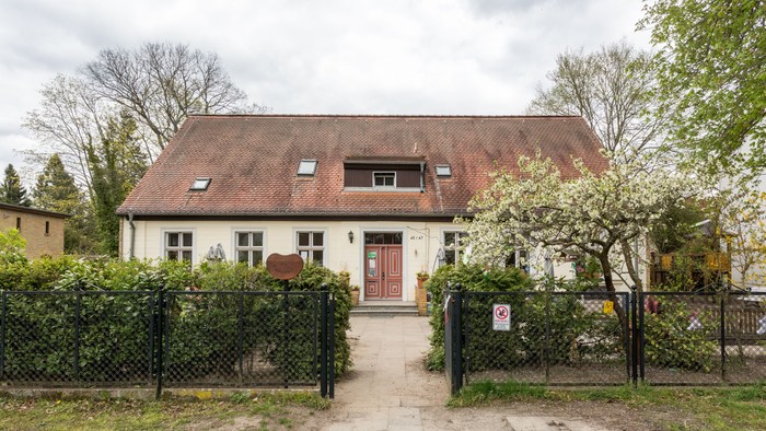Das Bild zeigt ein zweistöckiges Haus mit rotem Dach und weißer Fassade, umgeben von einem schwarzen Zaun und Büschen.