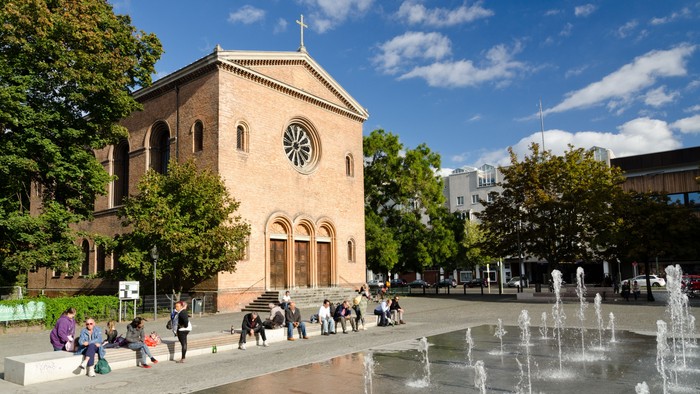 Ein Brunnen vor einem Kirchengebäude, Menschen sitzen und gehen umher