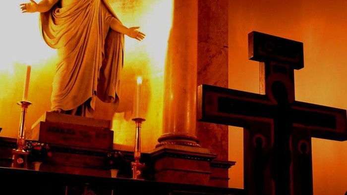 En statue af Jesus med udstrakt arme i en kirke med lysede lysstake og et stort kors.