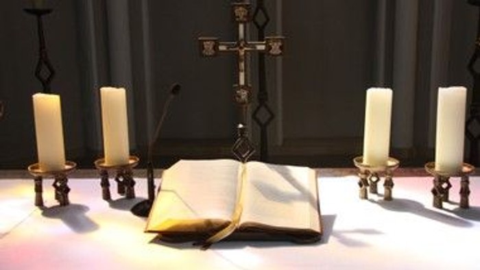 Religiöser Altarschmuck mit einem aufgeschlagenen Buch, vier Kerzen und einem dekorativen Kreuz.