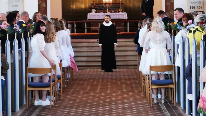 En præst står ved alteret i en kirke, omgivet af en forsamling.