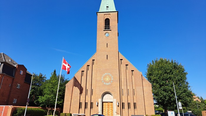 En murstenskirke med et højt spir, nationalflag og klar blå himmel, med en blå bil parkeret i nærheden.