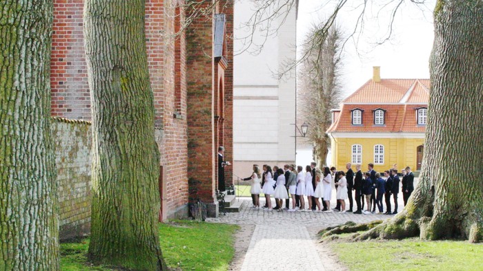 En gruppe mennesker i formelt tøj går langs en sti ved siden af en historisk murstensbygning og et gult hus.