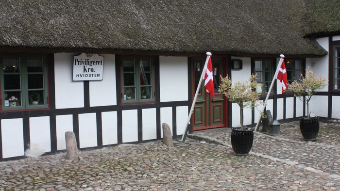 En hvidkalket hytte med sort træ og tagstroh, med to danske flag og to blomsterpletter ved indgangen