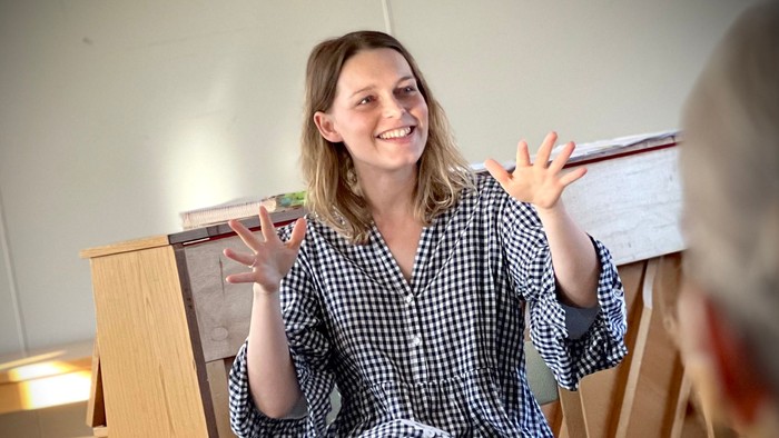 En kvinde sitter ved et piano og holder en flaske, mens hun smiler og vinkler.