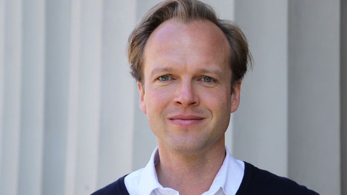 En mand med kort hår og blå øjne, der smiler og bærer en mørk pullover over et hvidt skjorte.