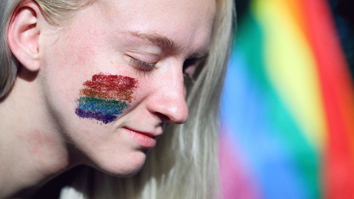 Eine junge Frau mit einem Regenbogen-Glitzer auf der Wange vor einem bunten Hintergrund.