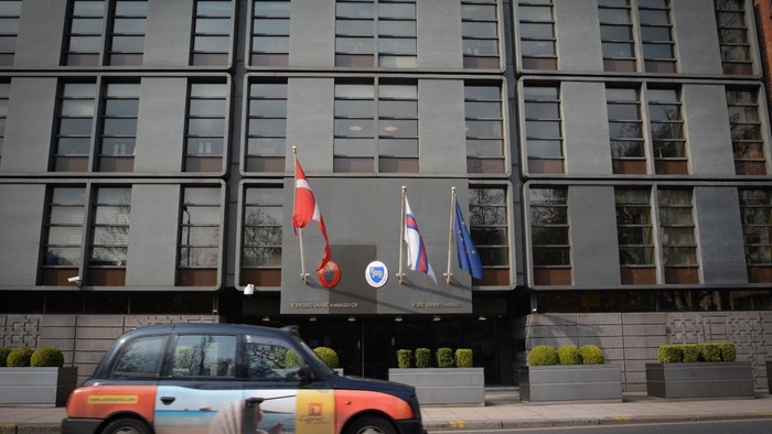 En rød taxi kører forbi et stort, graet kontorbygning med tre flagg.