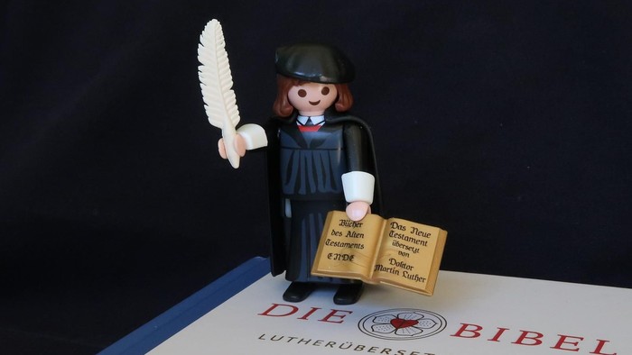 En lille figur med fjer og bog holdt op af en bog med tittelen "Die Bibel"