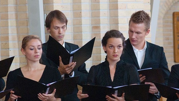 En gruppe af korister står i en hal og synger fra partiturer.