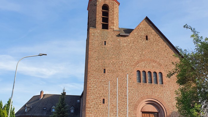 Ein Ziegelbau mit hohem Turm und Eingangstreppe, umgeben von Bäumen und einem klaren blauen Himmel.