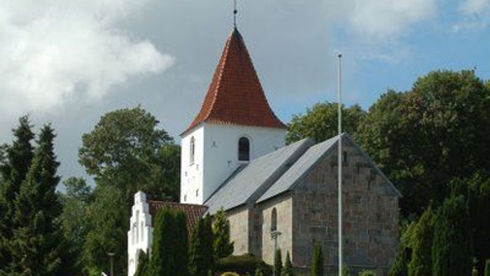 En lille kirke med rød spire og blå tag, omgivet af træer og en flagstang.